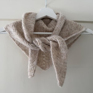 Könnte beinhalten: Ein beigefarbener Strickschal wird auf einem weißen Kleiderbügel präsentiert. Der Schal besteht aus einem strukturierten Garn und hat ein geknotetes Design auf der Vorderseite. Der Schal ist neutralfarben und wirkt weich und warm.