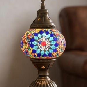 Puede incluir: Lámpara de mesa decorativa con un globo de cristal de mosaico en tonos azules, rojos, amarillos y verdes. La lámpara tiene una base de metal color bronce y un remate decorativo. La lámpara está enchufada a una toma de corriente.
