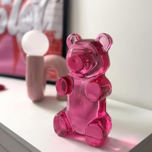 Op de afbeelding: Een transparante, felroze gummybeer-beeldje. De beer zit rechtop met zijn armen langs zijn zij. Het beeldje is gemaakt van glanzend materiaal en staat op een wit oppervlak. Een roze lamp en een ingelijste print staan op de achtergrond.