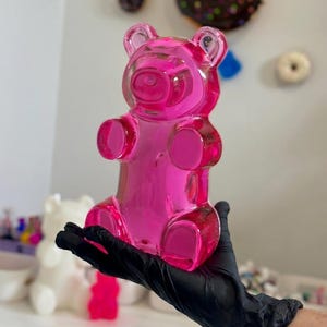 Peut inclure: Une grande figurine transparente en forme d'ours en gélatine rose vif. L'ours est tenu par une main gantée. L'ours a la forme classique d'un ours en gélatine avec des traits arrondis. La figurine est faite d'un matériau brillant et translucide.