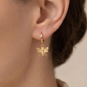 May include: A small gold hoop earring featuring a dangling bee charm. The bee charm is detailed with textured wings and a segmented body, suspended from a thin, polished gold hoop that secures through the earlobe with a simple closure.