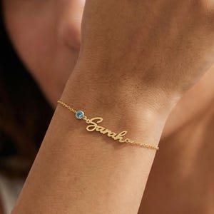 Könnte beinhalten: Ein zartes goldenes Armband mit dem Namen "Sarah" in Schreibschrift. Das Armband hat einen kleinen, runden, hellblauen Edelstein. Das Armband wird am Handgelenk getragen.