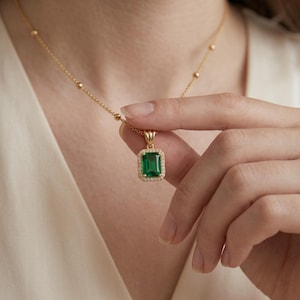 May include: A gold necklace featuring a rectangular emerald green gemstone pendant surrounded by a halo of small clear stones. The pendant hangs from a delicate gold chain with evenly spaced small gold beads along the length of the chain.