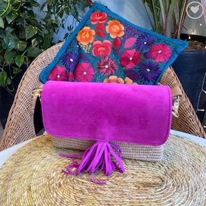 May include: A bright fuchsia suede handbag with a woven base and a long tassel. The bag features a gold-coloured clasp and a tan leather strap. An embroidered cushion with colourful floral designs is behind the bag.