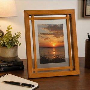 May include: A wooden picture frame with a sunset landscape photo. The frame is a warm, light brown color and has a rectangular shape with a unique design. The photo features a vibrant sunset over a body of water.