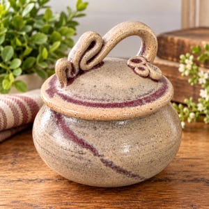 May include: A round, speckled ceramic jar with a matching lid featuring a curved handle and decorative clay accents. The piece has a light beige base color with dark maroon glaze streaks running across the lid and the body of the container.