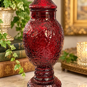 May include: A deep red glass apothecary jar with a textured, bumpy surface. The vessel features a wide, rounded body, a narrow neck, and a matching removable lid with a knobbed top. It stands on a circular, textured pedestal base.