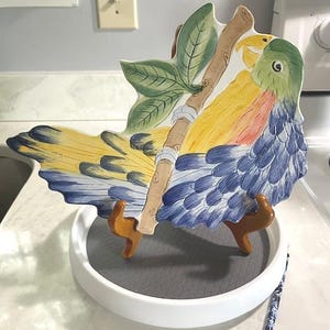 Puede incluir: Un plato decorativo de cerámica con forma de loro con colores amarillo, azul, verde y naranja. El plato está apoyado en un soporte blanco con patas de madera. El loro está posado en una rama marrón con hojas verdes.