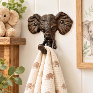 May include: A dark brown, textured cast iron wall hook shaped like an elephant head. The trunk curves upward to form the hook, which holds a cream-colored fabric towel featuring a repeating pattern of small, light brown elephant silhouettes.