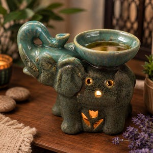 May include: A teal ceramic elephant oil burner featuring a glossy finish with dark speckles. The elephant has its trunk raised, and a circular bowl sits on its back. Cutout shapes on the body allow light to shine through from an internal source.