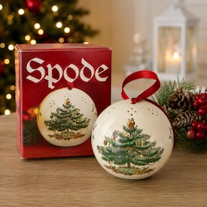 May include: A cream-colored ceramic pomander ball featuring a detailed Christmas tree illustration with a red ribbon hanger. The pomander is displayed next to its red cardboard box, which is printed with the brand name Spode in white text.