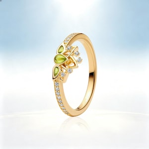 Puede incluir: Un anillo de oro con una banda incrustada de pequeñas piedras claras. La parte superior del anillo muestra un diseño decorativo con dos piedras de color verde claro en forma de pera y varias piedras claras pequeñas en un marco dorado.