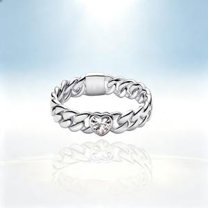 May include: A silver-toned chain link ring featuring a central heart-shaped clear gemstone. The band is constructed from interlocking metal links with a smooth, polished finish, and a flat rectangular metal segment is positioned at the top.