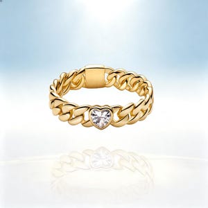 May include: A gold-toned chain link ring featuring a central heart-shaped clear gemstone. The band is constructed from interlocking metal links with a smooth, polished finish and a flat rectangular segment at the top opposite the heart stone.