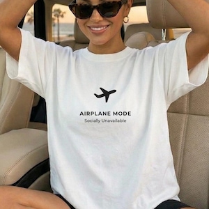 May include: A woman wearing a white t-shirt with the text "AIRPLANE MODE Socially Unavailable" and a black airplane graphic. She is wearing black shorts, white trainers, and sunglasses. The car interior is beige.