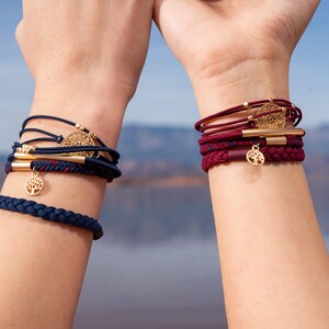 May include: Two wrists adorned with bracelets. One wrist features a navy blue braided bracelet and multiple navy blue and red cord bracelets with gold accents and a tree of life charm. The other wrist displays burgundy bracelets with gold accents and a tree of life charm.