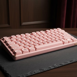 May include: A pink mechanical keyboard with rounded keycaps. The keyboard has a retro design with gold-coloured accents on the corners. The keys are light pink with white lettering. The keyboard is placed on a dark grey surface.