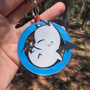Peut inclure: Un porte-clés circulaire représentant une silhouette blanche évoquant un fantôme avec des ailes de chauve-souris noires, insérée dans un anneau bleu vif. L'ensemble est fixé à un anneau métallique par une petite pièce de connexion rouge texturée.