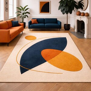 Peut inclure: Un tapis de sol crème avec un grand motif abstrait bleu marine et orange. Le tapis présente une forme incurvée bleu marine, un ovale orange et un petit cercle orange, tous reliés par un fin trait doré. Le tapis est dans un salon.
