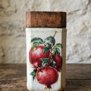 Op de afbeelding: Een rechthoekige keramische pot met een houten deksel. De pot is versierd met een levendige illustratie van rode granaatappels en groene bladeren. De pot staat op een houten oppervlak.