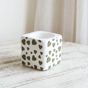 May include: A white, square-shaped planter with a textured surface. The planter is decorated with olive green heart-shaped patterns. The interior of the planter is hollow. The planter is sitting on a white wooden surface.