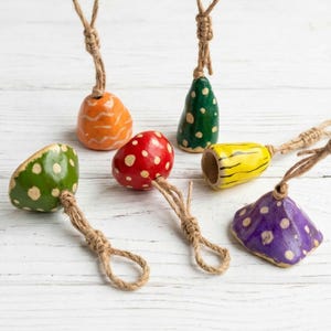 May include: A collection of colorful, hand-painted ceramic ornaments. The ornaments are in various shapes, including bell and mushroom designs, and feature polka dots. Each ornament has a jute string for hanging. Colors include orange, green, red, yellow, and purple.