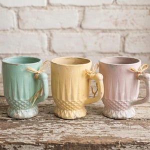 Puede incluir: Tres tazas decorativas de cerámica en colores pastel: verde menta, amarillo pálido y rosa suave. Cada taza presenta una superficie texturizada, un asa decorativa y un lazo de rafia. Las tazas se exhiben sobre una superficie de madera envejecida.