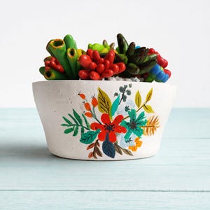 May include: A white ceramic planter with a hand-painted floral design, filled with colorful artificial succulents. The succulents are various shades of red, green, and blue. The planter sits on a light blue surface.