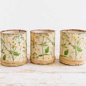 Könnte beinhalten: Drei zylindrische Behälter mit floralen Mustern. Die Behälter bestehen aus wiederverwendeten Dosen, die mit gelben und grünen botanischen Illustrationen verziert sind. Jeder Behälter ist am Boden mit einem natürlichen Bindfadenband umwickelt, was einen rustikalen Touch verleiht.