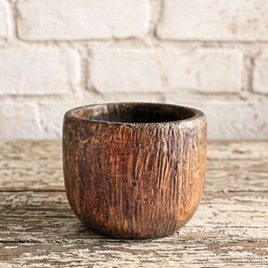 May include: A small, handcrafted wooden bowl with a dark brown finish. The bowl has a rounded shape and a textured surface, showcasing the natural grain of the wood. The interior is dark, contrasting with the lighter exterior. The bowl sits on a distressed wooden surface.