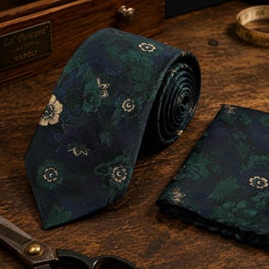 Può includere: Una cravatta blu navy scuro e una pochette abbinata con un motivo floreale ripetuto nelle tonalità del verde bosco, verde acqua e crema. Il tessuto ha una lucentezza sottile e i motivi floreali sono disposti su tutta la superficie.