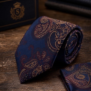 Può includere: Una cravatta blu navy caratterizzata da un dettagliato motivo paisley in oro e rosso scuro. La cravatta è piegata e accompagnata da una pochette abbinata, entrambe mostrano intricati motivi a vortice sulla superficie del tessuto.