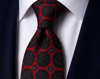 Puede incluir: Una corbata roja con un patrón repetitivo de medallones circulares de color azul marino oscuro con centros florales complejos. La corbata está anudada sobre una camisa blanca de cuello rígido, bajo una chaqueta de traje azul marino.