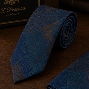 Può includere: Una cravatta arrotolata caratterizzata da un motivo intrecciato blu profondo e marrone con intricati disegni a vortice. La cravatta è posizionata vicino a una scatola scura con il testo L. Pescara stampato in oro. È visibile una pochette abbinata.