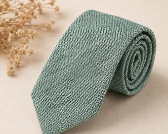 Può includere: Una cravatta arrotolata caratterizzata da una trama testurizzata in un colore verde salvia solido. Il tessuto presenta un visibile motivo a tratteggio incrociato con sottili variazioni di tono, creando un aspetto uniforme sulla cravatta.