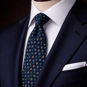 May include: A dark navy blue blazer worn over a crisp white dress shirt with a dark navy blue necktie featuring a repeating pattern of small teal and gold floral motifs and a white pocket square tucked into the blazer breast pocket.