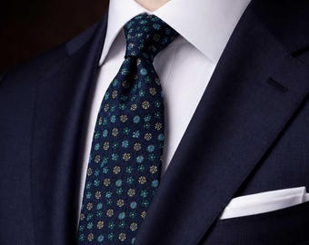 Puede incluir: Una americana azul marino oscuro sobre una camisa de vestir blanca impecable con una corbata azul marino oscuro que presenta un patrón repetitivo de pequeñas flores en tonos azul cerceta y dorado y un pañuelo blanco en el bolsillo.