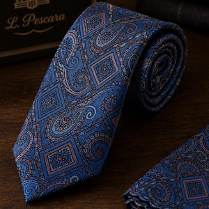 Può includere: Una cravatta blu con un motivo ripetuto di rombi e volute paisley nelle tonalità dell'azzurro, blu scuro e rosa tenue. Il tessuto presenta una visibile trama diagonale. Una pochette abbinata è parzialmente visibile nell'angolo.