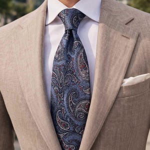 Op de afbeelding: Een marineblauwe stropdas met paisley-patroon en rode en witte accenten is geknoopt over een wit overhemd. De stropdas wordt gecombineerd met een beige blazer en een witte pochet is zichtbaar.