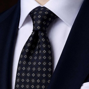 Puede incluir: Una corbata azul marino oscuro con un patrón geométrico repetitivo de pequeños rombos blancos y gris claro. La corbata está anudada sobre una camisa de vestir blanca de cuello rígido, que se lleva debajo de una chaqueta de traje azul marino.