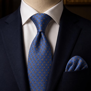 Pode incluir: Um paletó azul-marinho sobre uma camisa branca de colarinho, apresentando uma gravata azul com um padrão repetitivo de quadrados dourados e um lenço de bolso a condizer na lapela, criando um conjunto formal e coordenado.