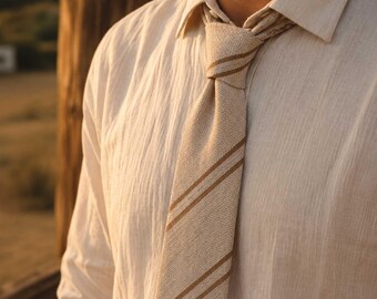 Può includere: Una cravatta beige testurizzata con due strisce diagonali marroni, indossata con una camicia abbottonata beige abbinata. La cravatta è annodata al colletto, mostrando una struttura in tessuto intrecciato con un motivo visibile e coerente.