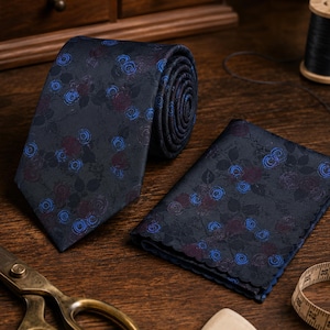 Peut inclure: Une cravate bleu marine foncé et une pochette de costume assortie, toutes deux ornées d'un motif répétitif de petites roses bleues et violet foncé avec des feuilles. La pochette présente un bord festonné décoratif sur son pourtour.