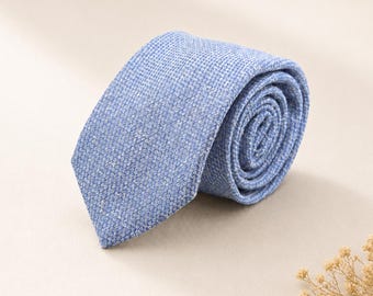 Può includere: Una cravatta arrotolata di colore azzurro caratterizzata da un motivo fine, testurizzato e maculato su tutto il tessuto. La cravatta è piegata in forma circolare, mostrando l'estremità a punta e l'intreccio complesso del materiale blu e bianco.