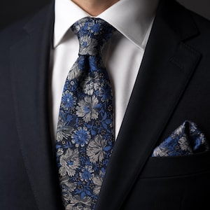 Op de afbeelding: Een stropdas en bijpassend pochet met een donkerblauwe achtergrond en een dicht, ingewikkeld bloemenpatroon in tinten blauw, grijs en wit. De set wordt gedragen met een wit overhemd en een effen donkerblauw colbert van een pak.