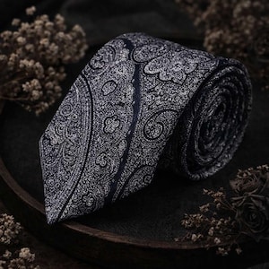 May include: A rolled navy blue tie with a white paisley pattern. The tie is made of a textured fabric and is displayed on a dark surface with dried flowers.