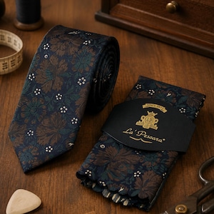 Könnte beinhalten: Eine dunkelmarineblaue Krawatte und ein passendes gefaltetes Einstecktuch mit einem braun-türkisfarbenen Blumenmuster und kleinen weißen Akzenten. Das Einstecktuch ist mit einer schwarzen Papierbanderole mit der Aufschrift La' Pescara versehen.