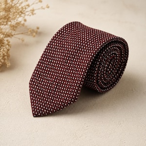 May include: A necktie in a deep burgundy red color featuring a repeating pattern of tiny white cross symbols. The tie is rolled up at one end, showing the textured fabric construction and the pointed tip of the blade in a clear, focused view.