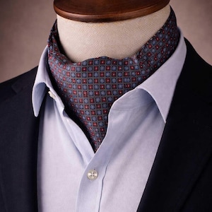 Può includere: Un ascot fantasia nei toni del grigio, rosso e blu, indossato con una camicia azzurra e un blazer blu navy. L'ascot presenta un motivo geometrico ripetuto. La camicia è abbottonata al colletto.