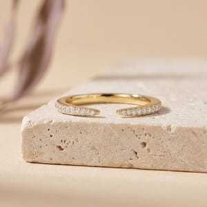Puede incluir: Un anillo abierto de oro con una banda pulida engastada con una serie de pequeñas piedras transparentes de corte redondo a lo largo del arco frontal. El anillo tiene una abertura en la parte superior, creando una forma de herradura.